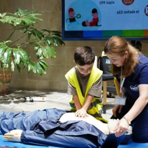 A young boy learning CPR from an instructor on a dummy indoors, with educational materials displayed.
