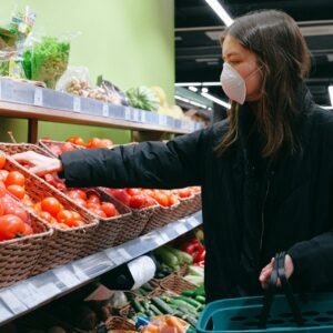 A woman wearing a mask selects fresh tomatoes in a grocery store aisle.